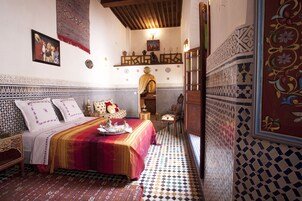 Quarto tradicional marroquino com piso estampado, cama com colcha vermelha e dourada e um tecido decorativo na parede.