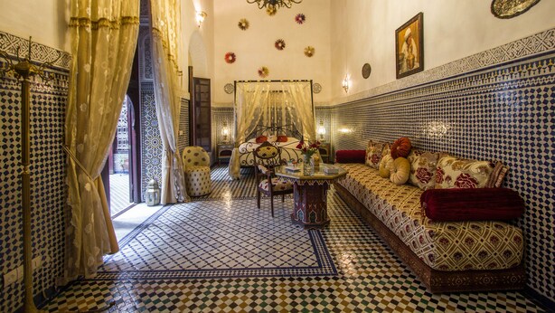Um quarto com piso xadrez, um sofá com almofadas estampadas, uma cama com dossel e um candelabro.