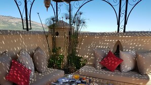 Terraço com sofá e mesa posta para o café da manhã, rodeado por plantas decorativas e vista para as colinas.
