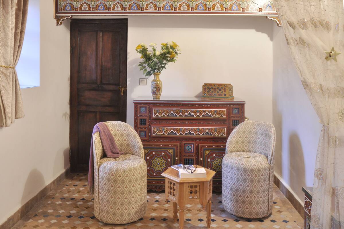 A room with two patterned armchairs, a wooden cabinet with decorative tiles, and a vase with flowers on the cabinet.