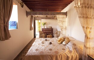 Quarto com cama grande, duas cadeiras, uma mesinha e janela com cortinas.