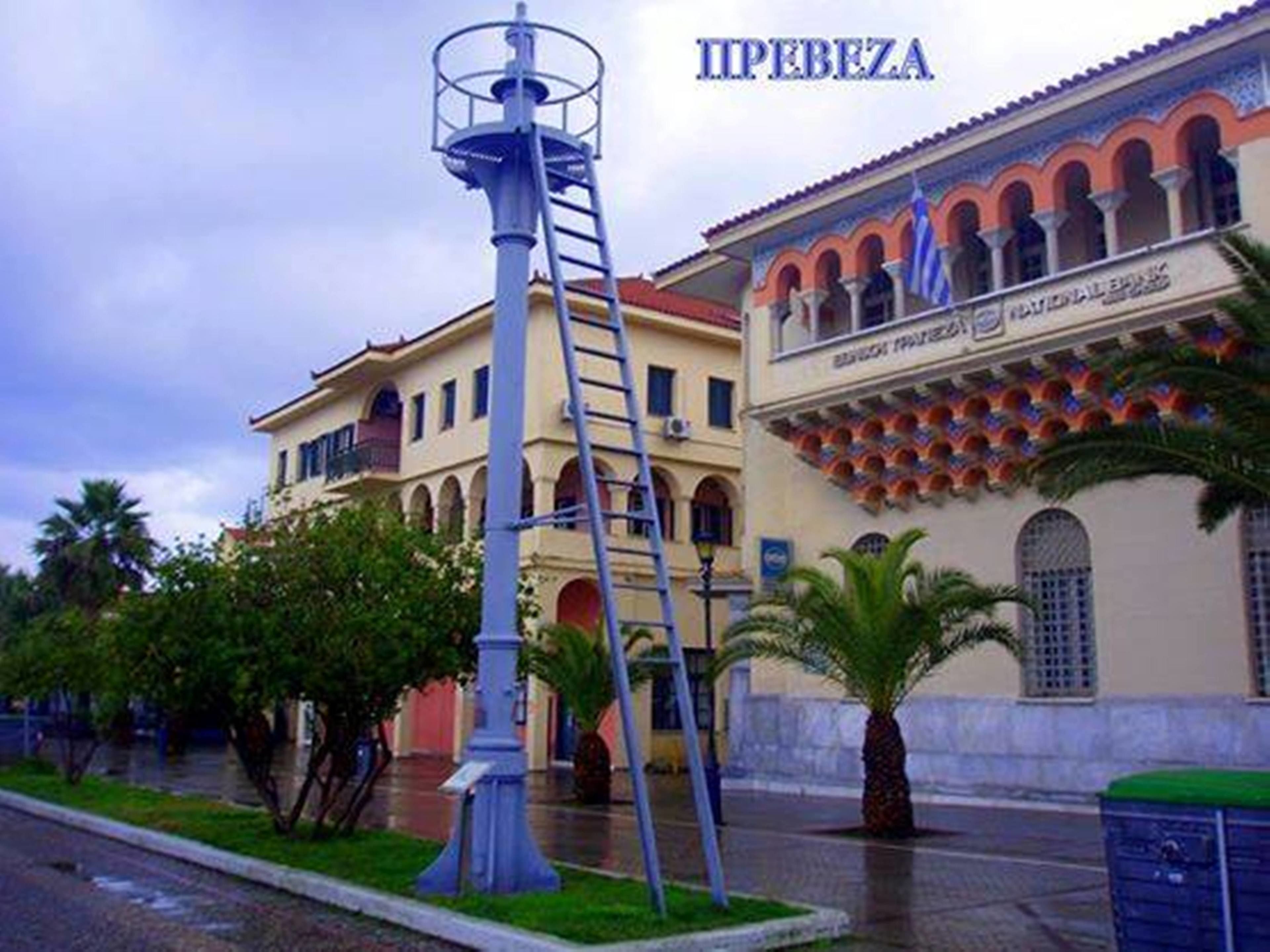 Une scène de rue avec une haute structure étroite en forme de tour, un bâtiment doté d’arcades et d’une enseigne indiquant « PIRBEZA », ainsi qu’un palmier.
