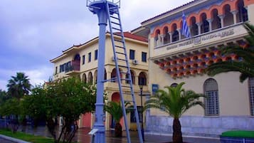 Une scène de rue avec une haute structure étroite en forme de tour, un bâtiment doté d’arcades et d’une enseigne indiquant « PIRBEZA », ainsi qu’un palmier.