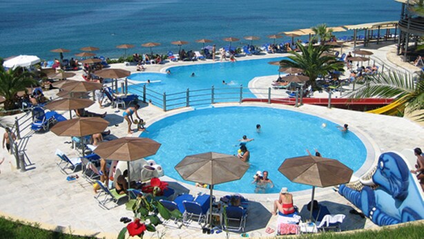 Una gran piscina al aire libre con varias pistas de natación, rodeada de sillas de descanso y sombrillas, con vistas a una playa.