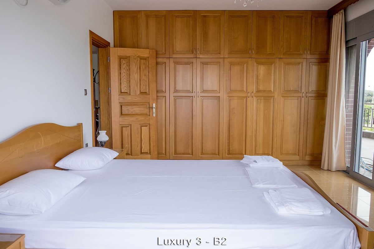 A large bed with white linens, a wooden headboard, and a view of a wooden wardrobe and balcony.