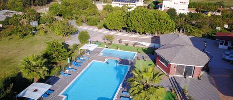 A large swimming pool with sun loungers and a covered area, surrounded by greenery and residential buildings.