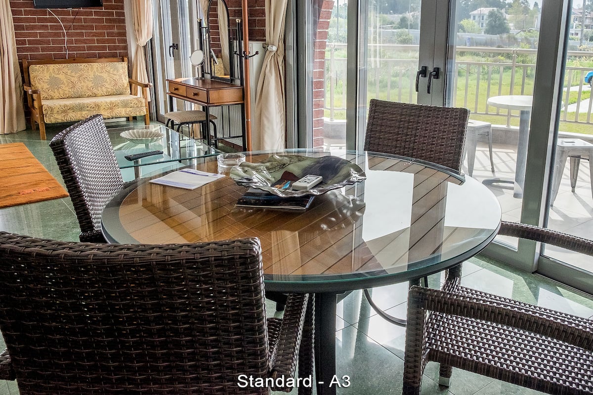 A round glass table with wicker chairs, a sofa, and a small table with a mirror and a lamp.