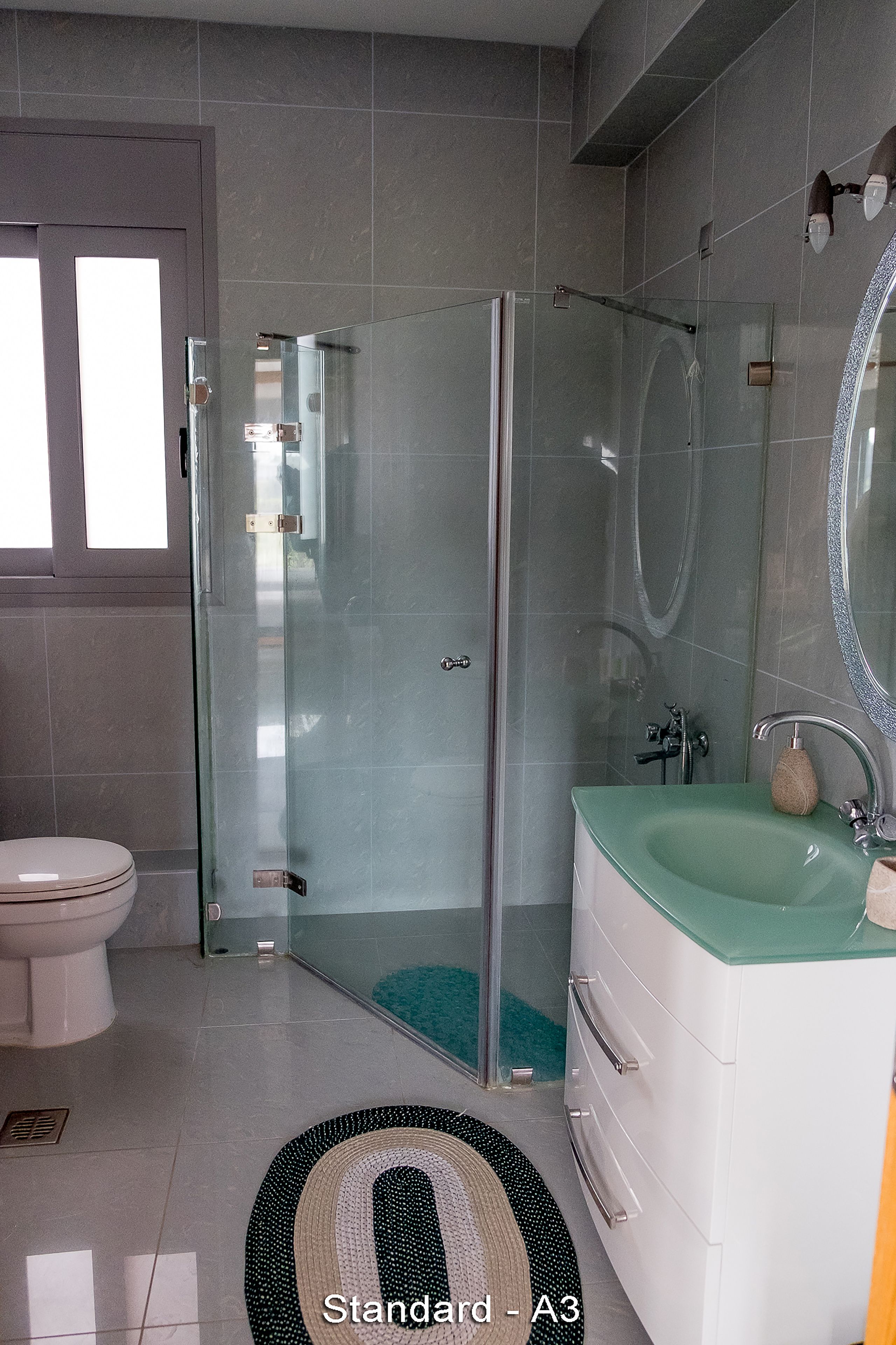 Une salle de bain moderne avec une douche à parois de verre, des toilettes blanches, un lavabo vert et un miroir doté d’un cadre décoratif.