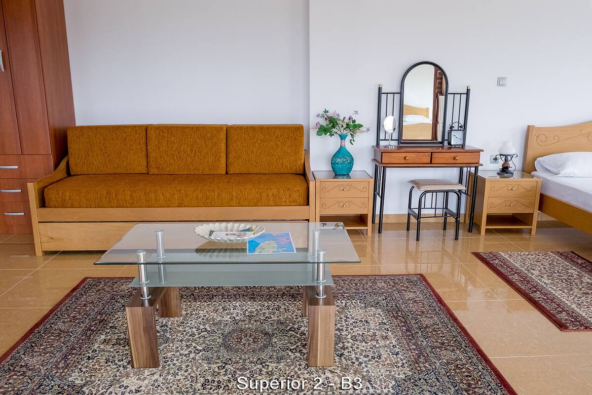 A room with a sofa, a glass-top coffee table, a wooden dresser, a mirror, a bedside table, and a bed.