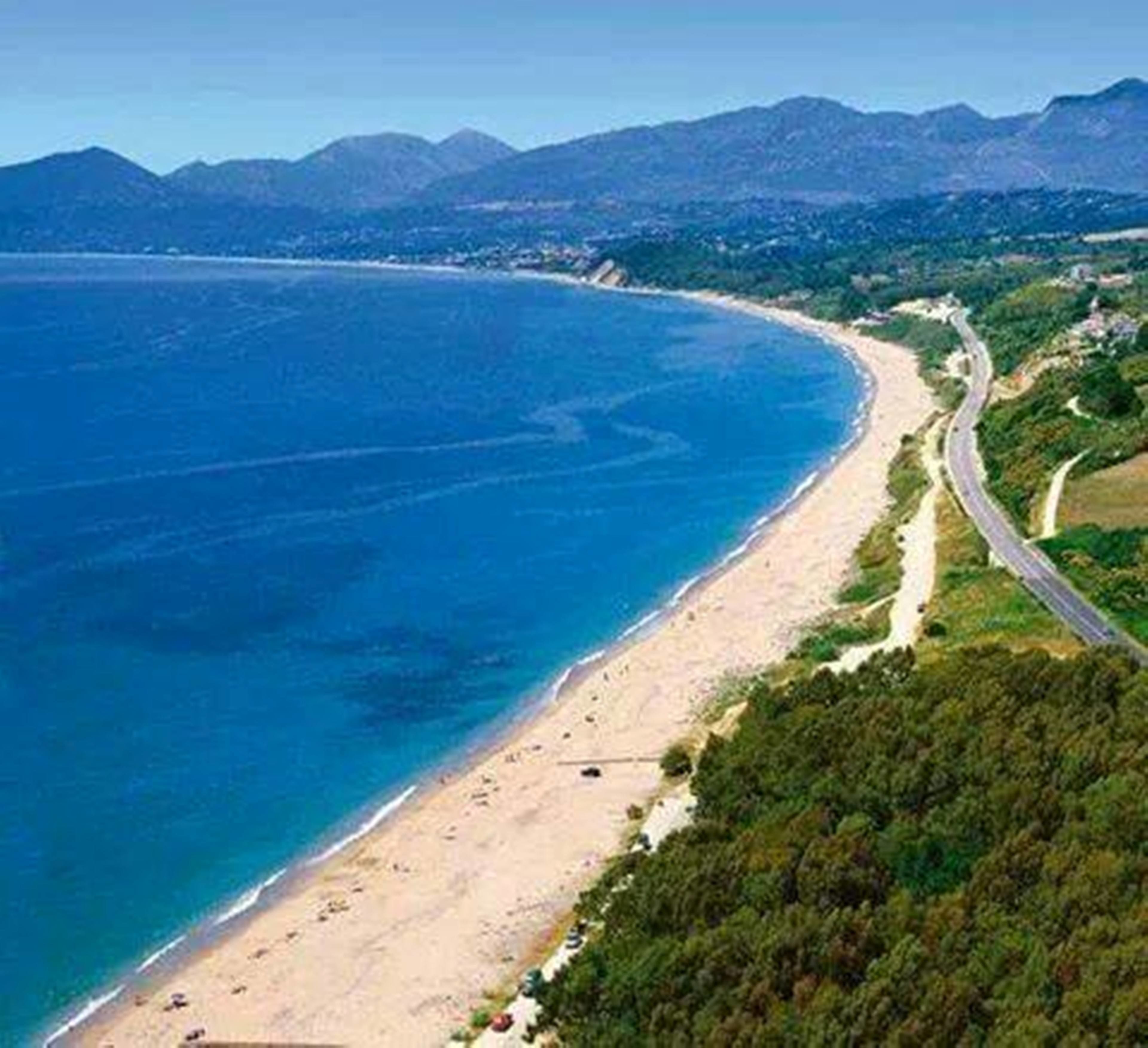 Luchtbeeld van een kustgebied met een zandstrand, een kronkelende weg en een bergketen op de achtergrond.