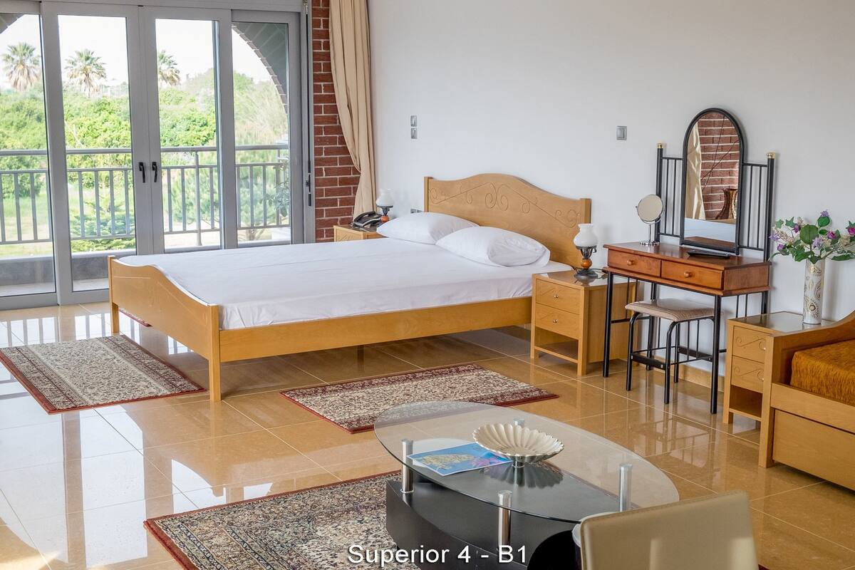 A well-lit hotel room with a bed, desk, and chair, a small table with a decorative item, and a view of greenery through large windows.