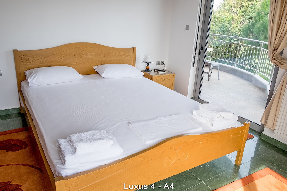 A Luxus 4 - A4 room with a wooden bed, white linens, a bedside table with a lamp, and a view of a balcony with greenery.