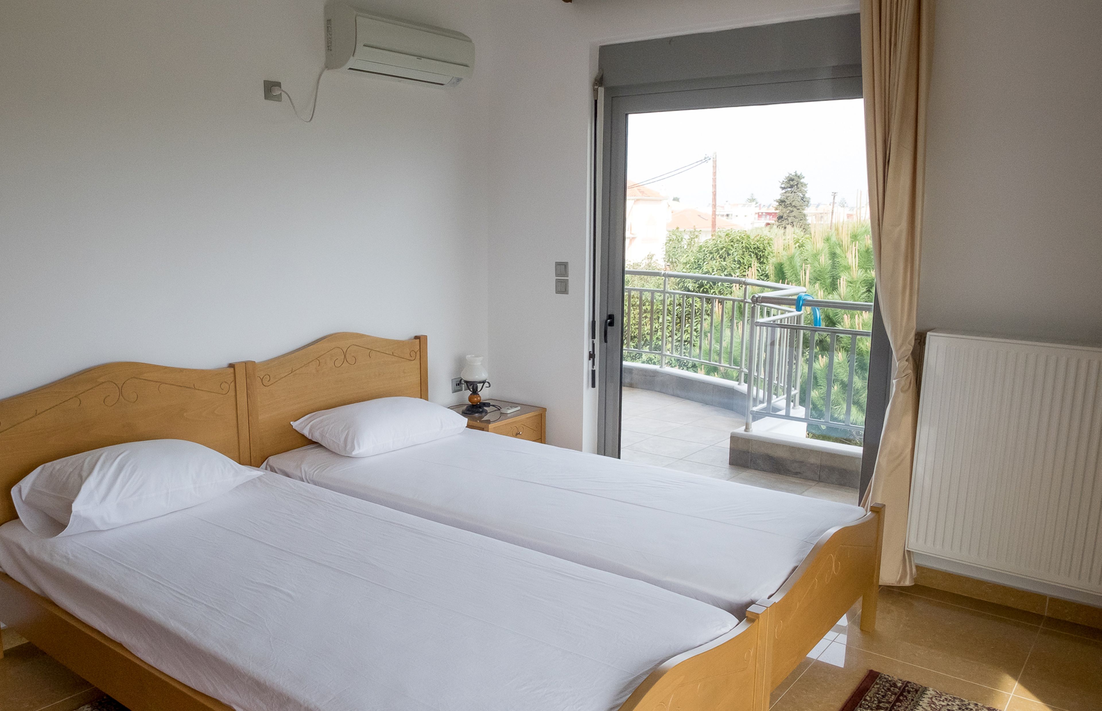 Un lit double avec du linge de lit blanc, une tête de lit en bois, une table de chevet avec une lampe, et une vue sur un balcon avec une balustrade.