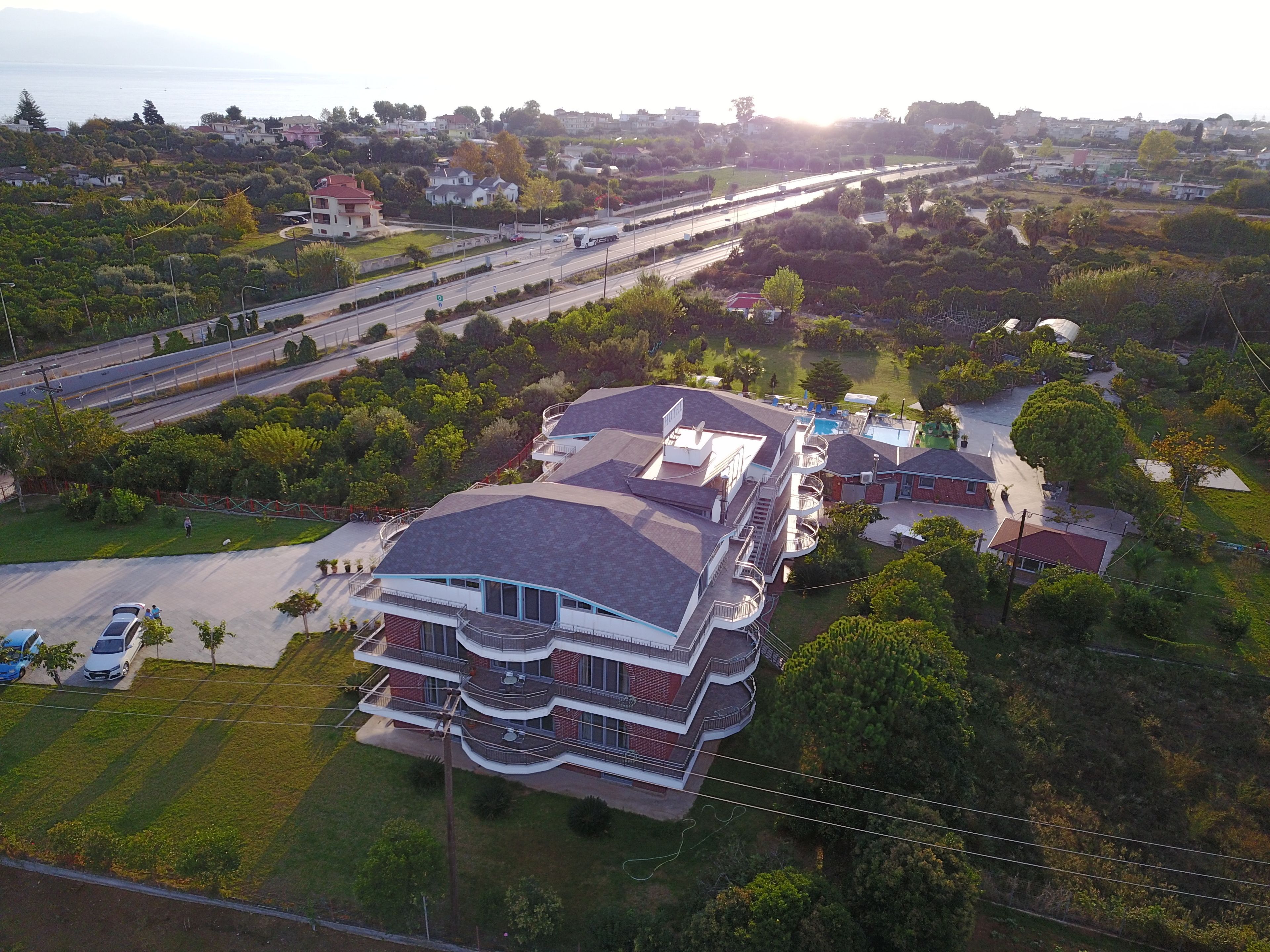 A multi-story building with balconies, surrounded by greenery and a road with vehicles.