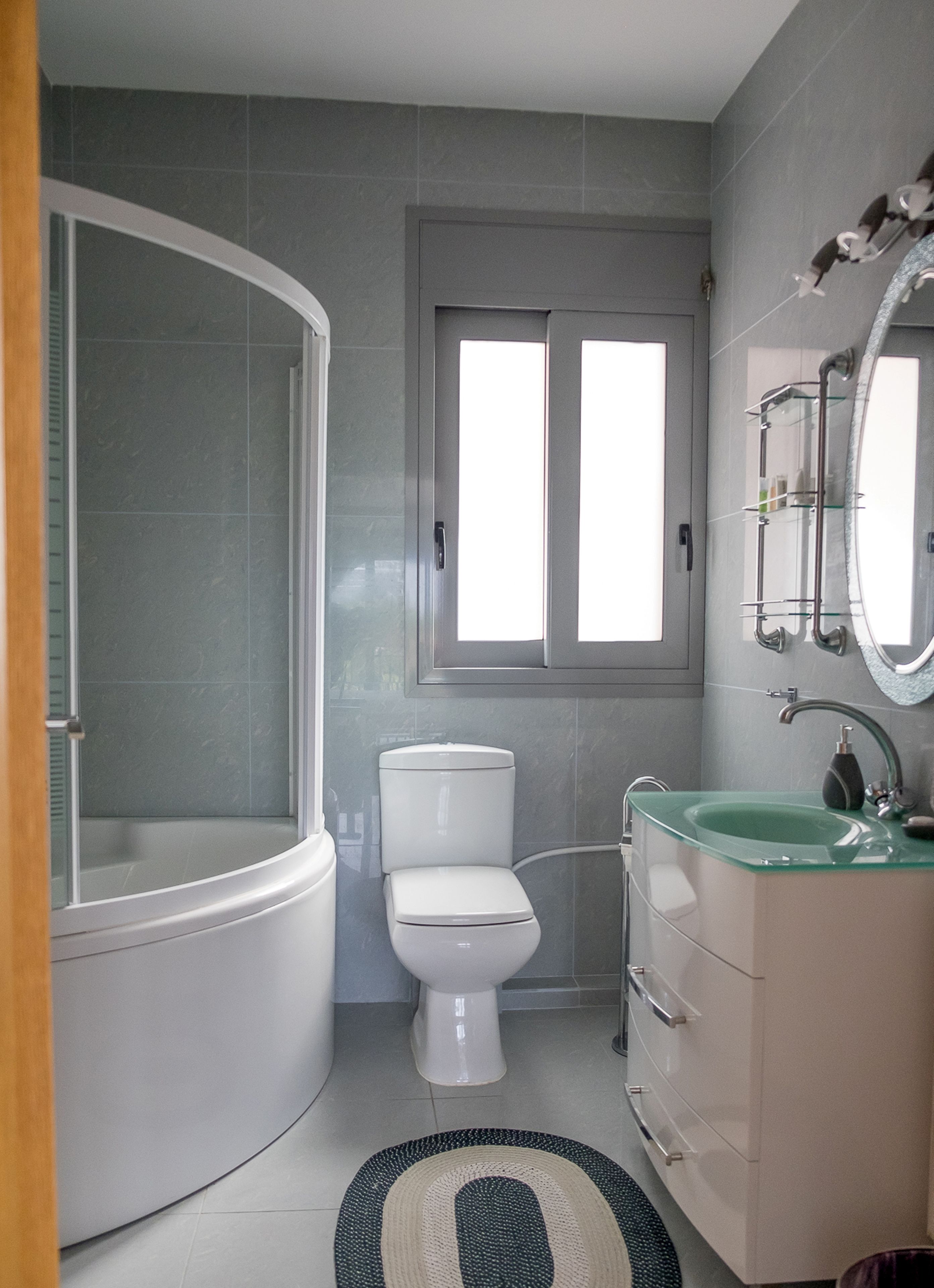 Un baño moderno con cabina de ducha de vidrio, inodoro, lavamanos con espejo y ventana sobre el inodoro.
