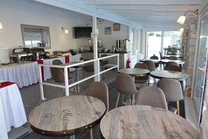 A restaurant with round wooden tables, white tablecloths, and a buffet area with various food items.