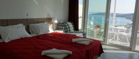 Habitación de hotel con una cama grande, una mesita de noche con lámpara, un otoman con estampado de cuadros y un balcón con vista al mar.
