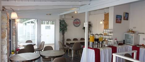 Un restaurant avec des tables et des chaises rondes, un espace buffet proposant des plats et des boissons, et une vue sur l’extérieur.
