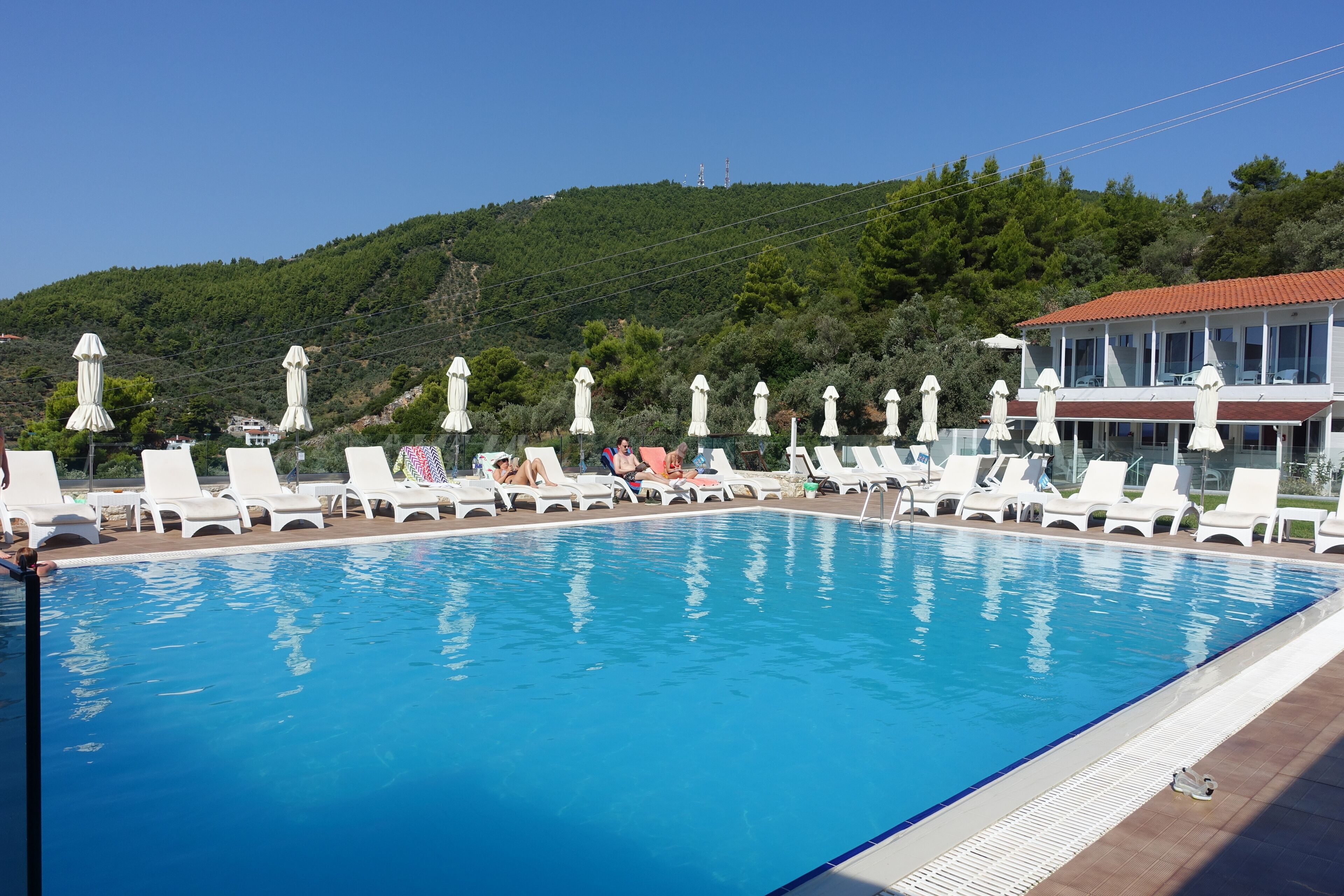 A swimming pool with white lounge chairs and umbrellas, surrounded by a building with a red-tiled roof.