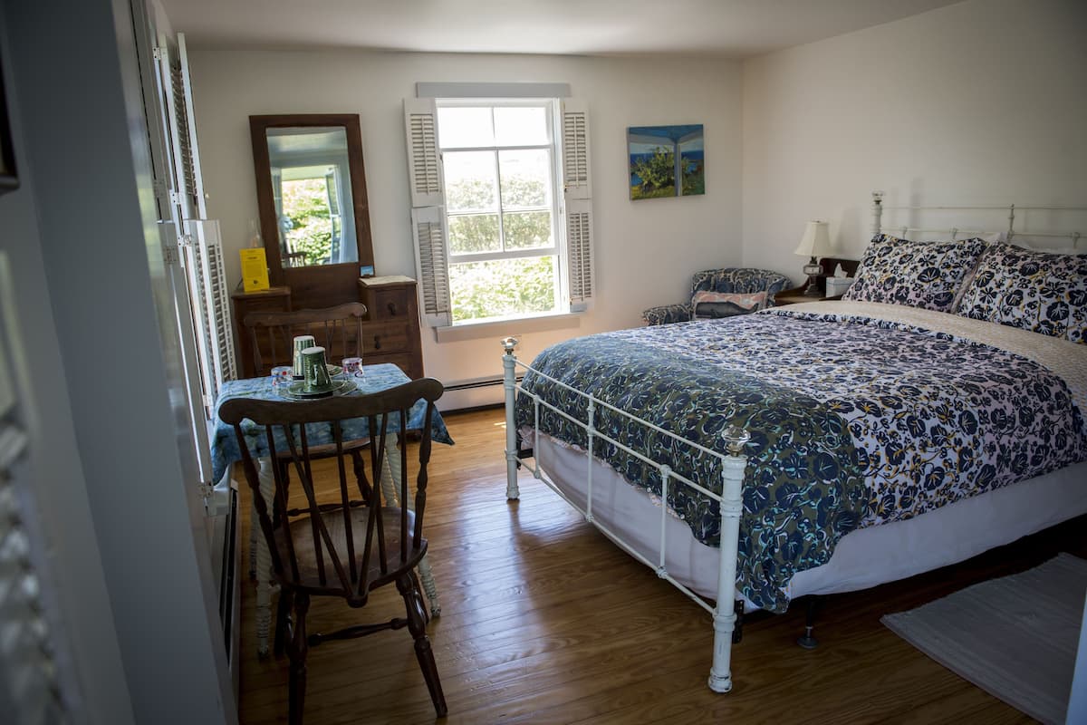 Una camera da letto con un letto, una sedia, un tavolo, una cassettiera, una finestra e un quadro appeso al muro.