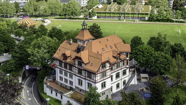 A traditional half-timbered building with a prominent tower, situated near a sports field and a road. - Hotel Militärkantine (St. Gallen)