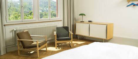 A hotel room with a bed, two chairs, a radiator, a window with a view of greenery, and a cabinet with a lamp.