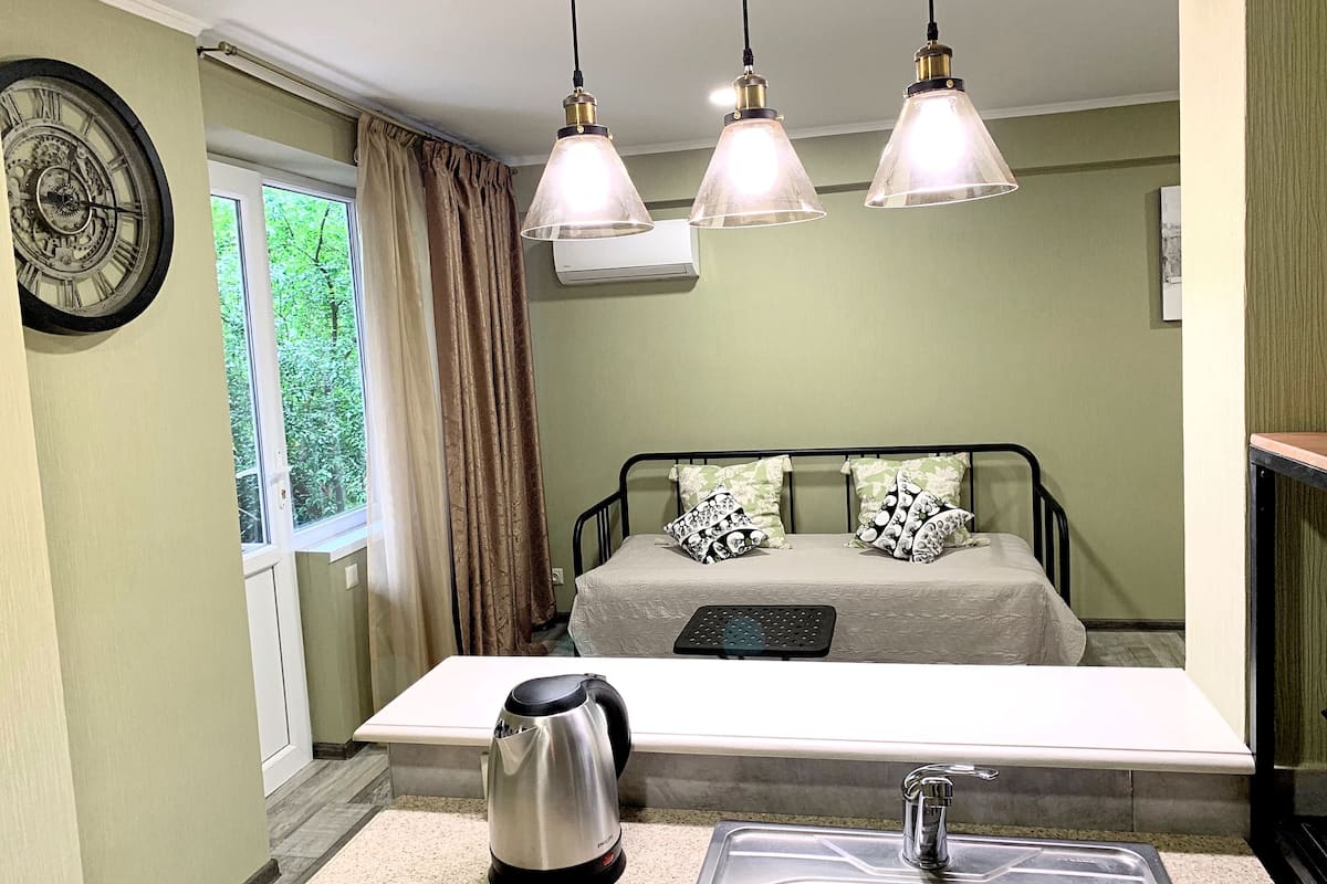 A bedroom with a bed, a bedside table, a kettle, a sink, and a window with curtains.