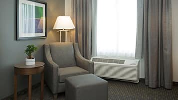 A hotel room with a grey armchair, a small wooden side table with a potted plant, a standing lamp, and a window with curtains.