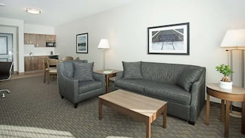 Una habitación de hotel moderna con un sofá de cuero gris, una mesa de centro de madera, una mesita auxiliar pequeña, una lámpara y un cuadro enmarcado en la pared.