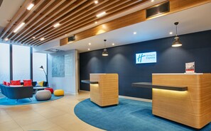 A modern reception area with wooden reception desks, a blue carpet, and a large window providing natural light. - Holiday Inn Express London - Ealing by IHG (London)