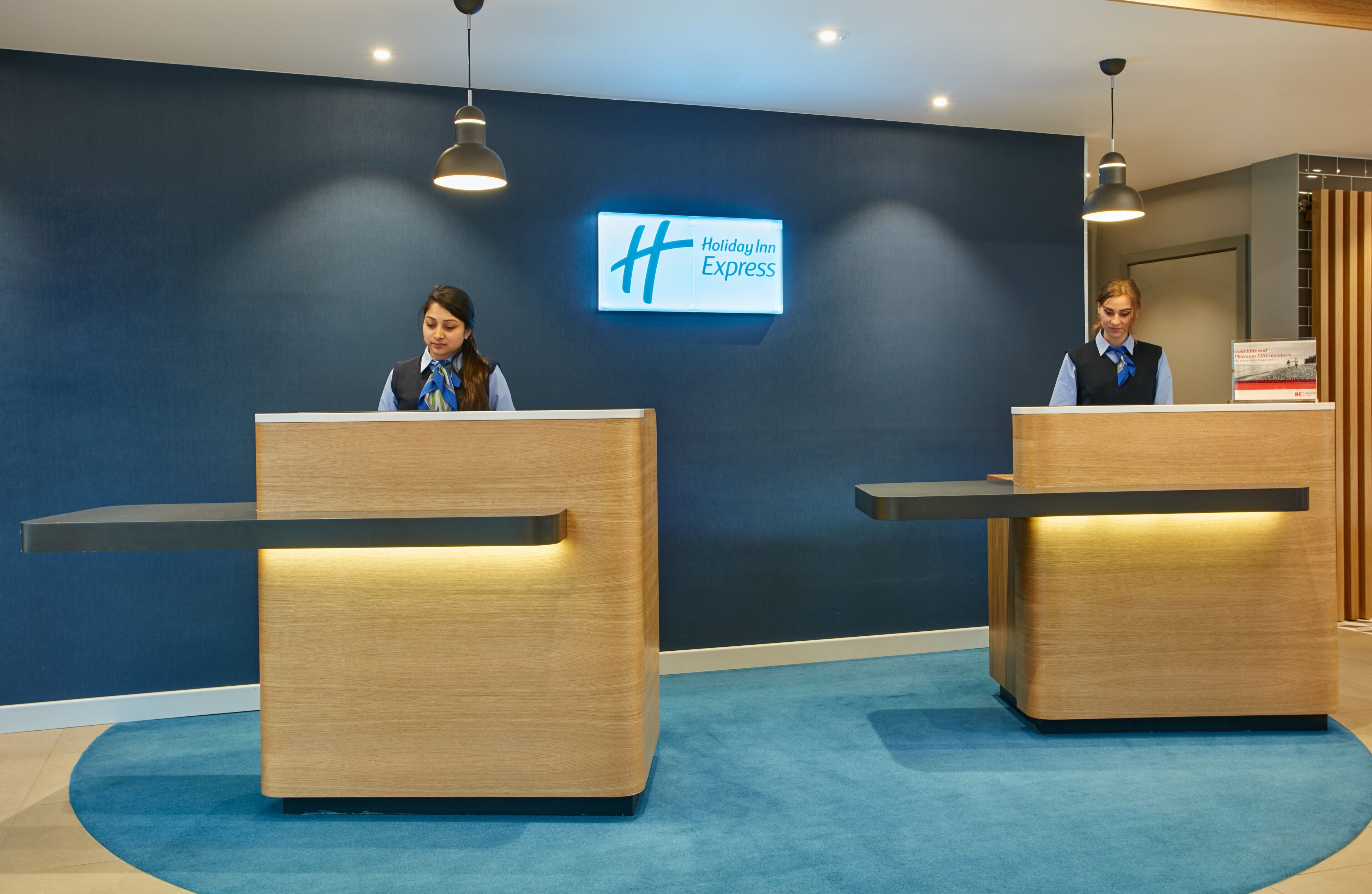 A reception desk area with two receptionists, a blue wall with a Holiday Inn Express sign, and a blue carpet.