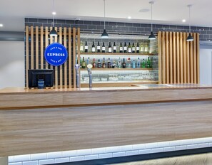 A modern bar area with a wooden counter, a blue sign indicating it's a cafe and bar, and shelves stocked with various bottles.