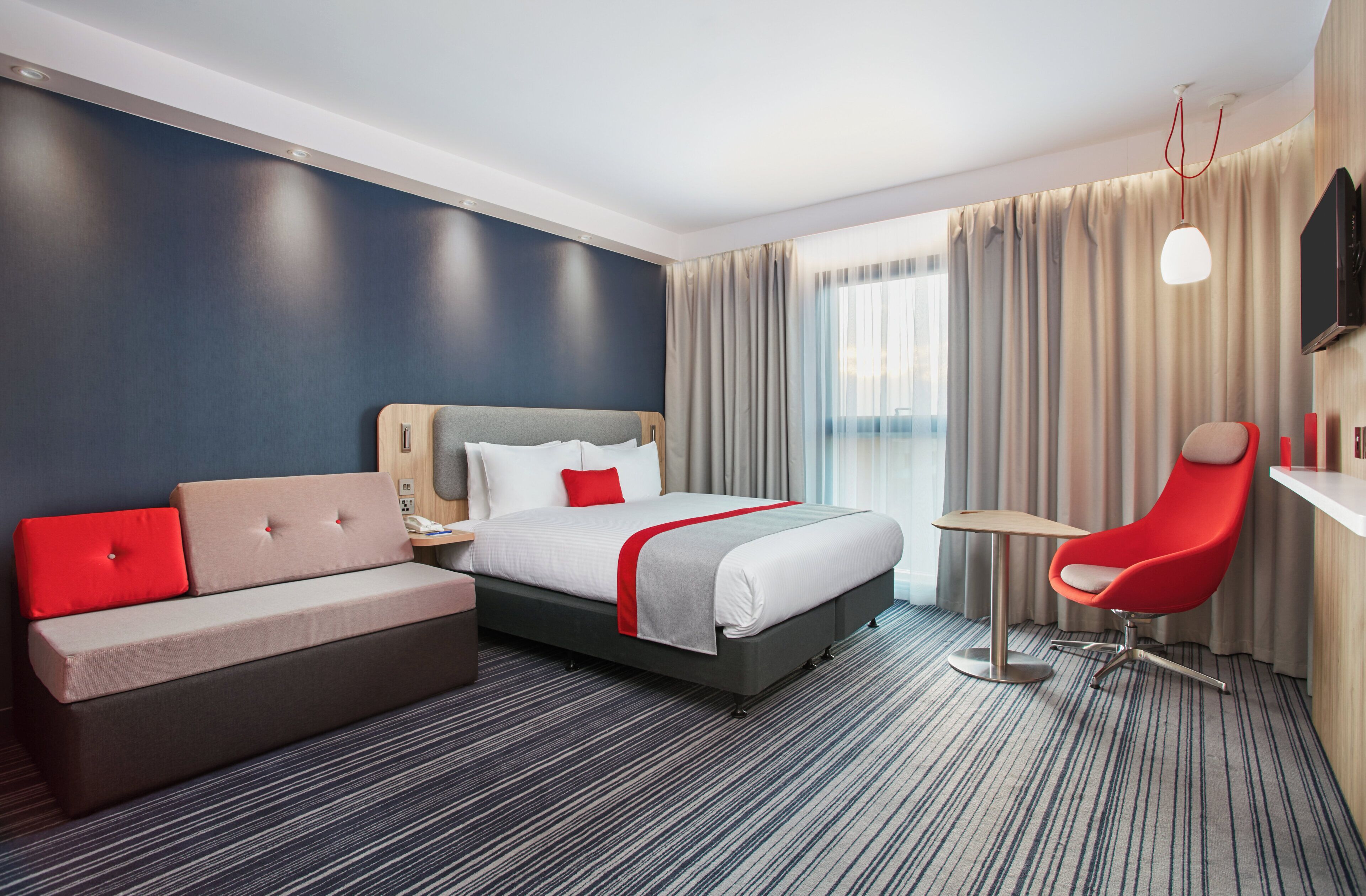 A hotel room with a bed, a sofa, a desk, a chair, and a TV.