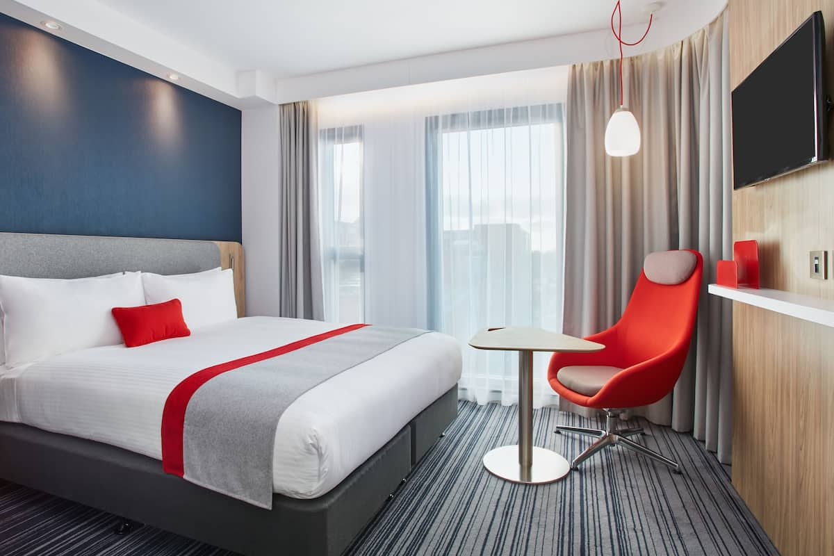 Een hotelkamer met een groot bed, een rode stoel, een tafeltje, een televisie en uitzicht over de stad.