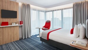 A hotel room with a bed, a red chair, a small table, a TV, and a city view. - Holiday Inn Express London - Ealing by IHG (London)