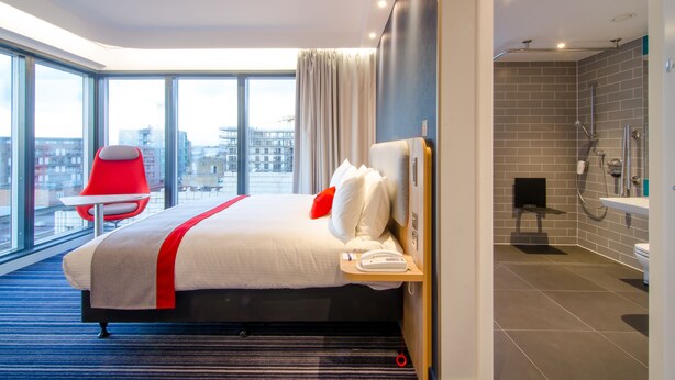 A modern hotel room with a large bed, a red chair, a view of the city, and a separate bathroom with a shower and sink. - Holiday Inn Express London - Ealing by IHG (London)