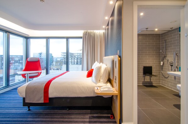 A modern hotel room with a large bed, a red chair, a view of the city, and a separate bathroom with a shower and sink. - Holiday Inn Express London - Ealing by IHG (London)