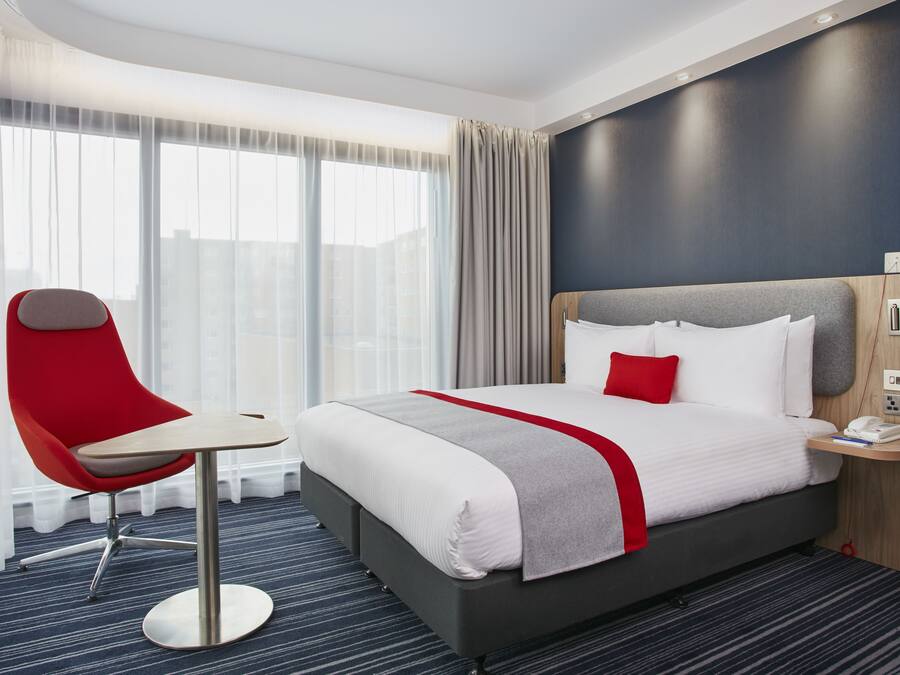 A hotel room with a large bed, a red chair, a small table, and a nightstand with a phone and a lamp.