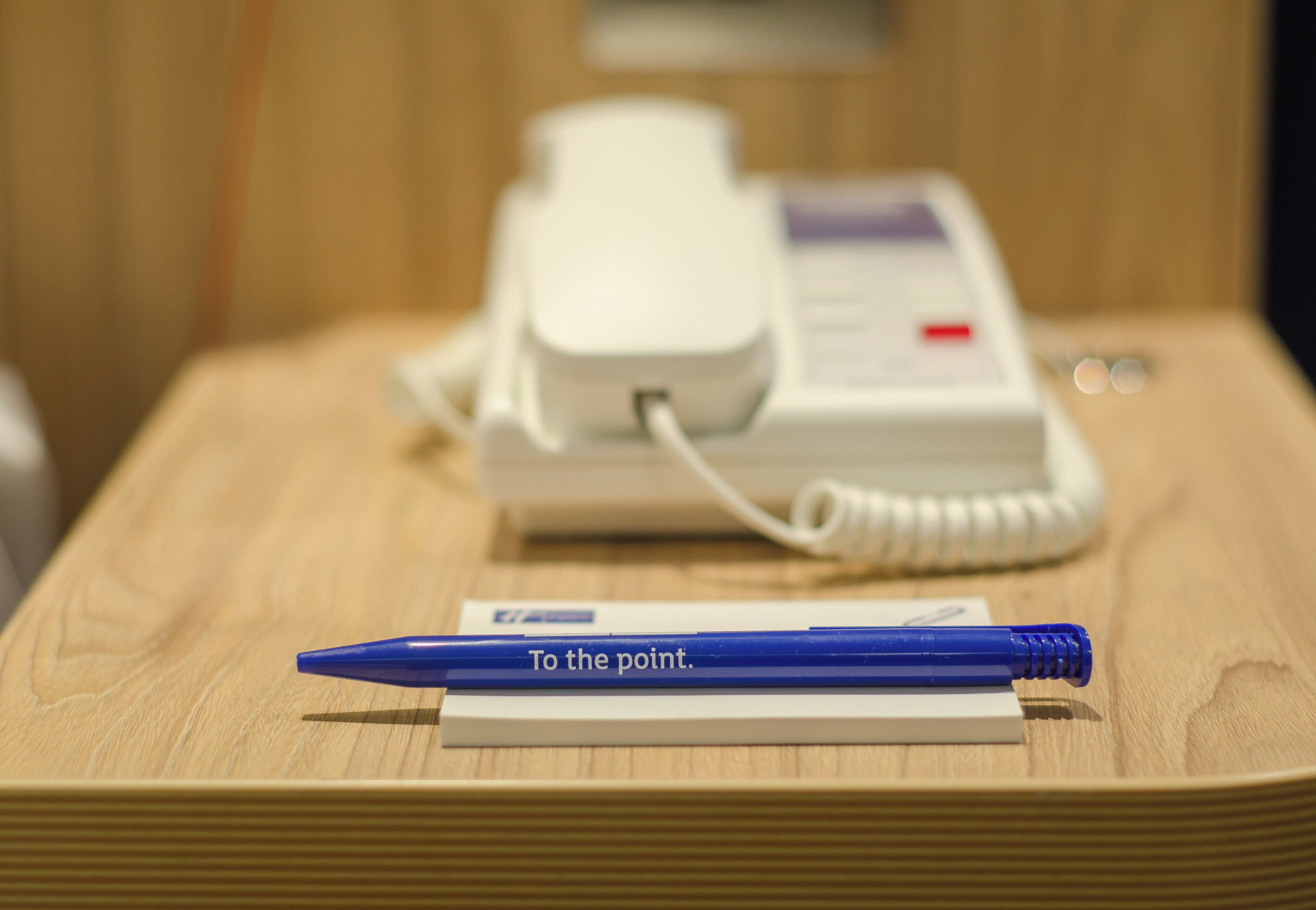 A wooden table with a white telephone and a blue pen on a piece of paper.