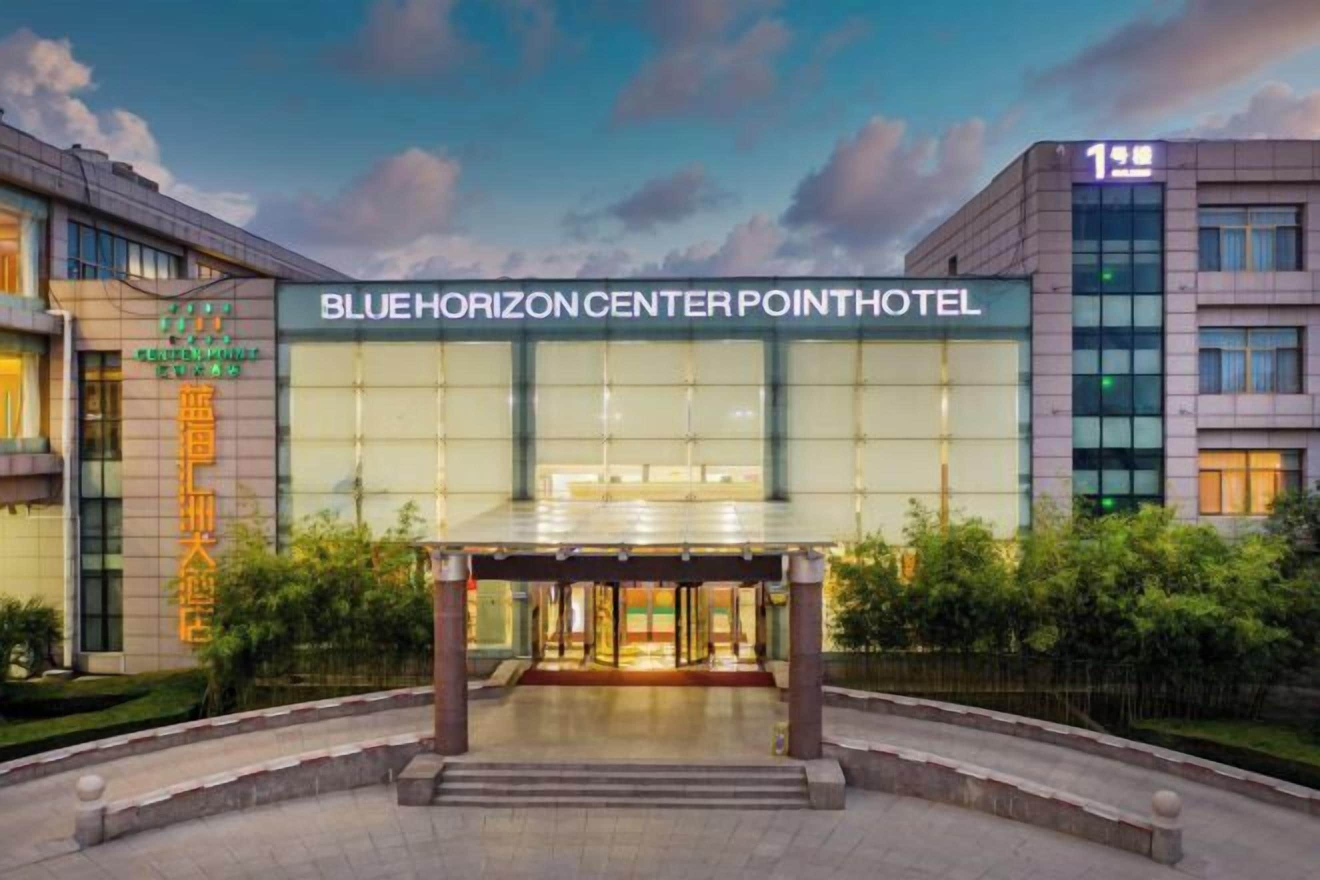 The exterior of the Blue Horizon Center Point Hotel with a modern glass facade, a covered entrance, and a landscaped area in front.