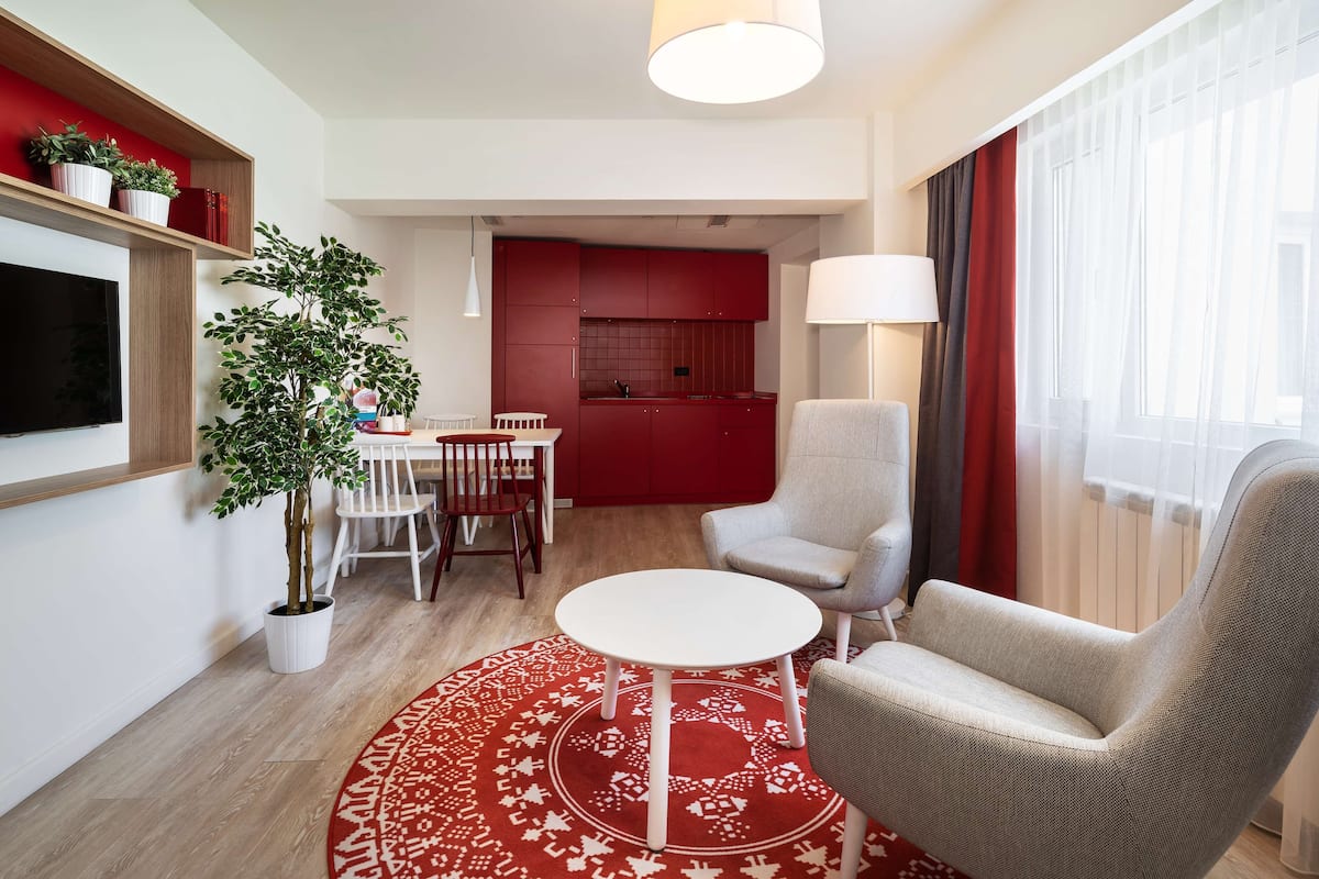 A modern living room with a round coffee table, a grey armchair, a TV mounted on the wall, a dining area with a red table, and a kitchen in the background.
