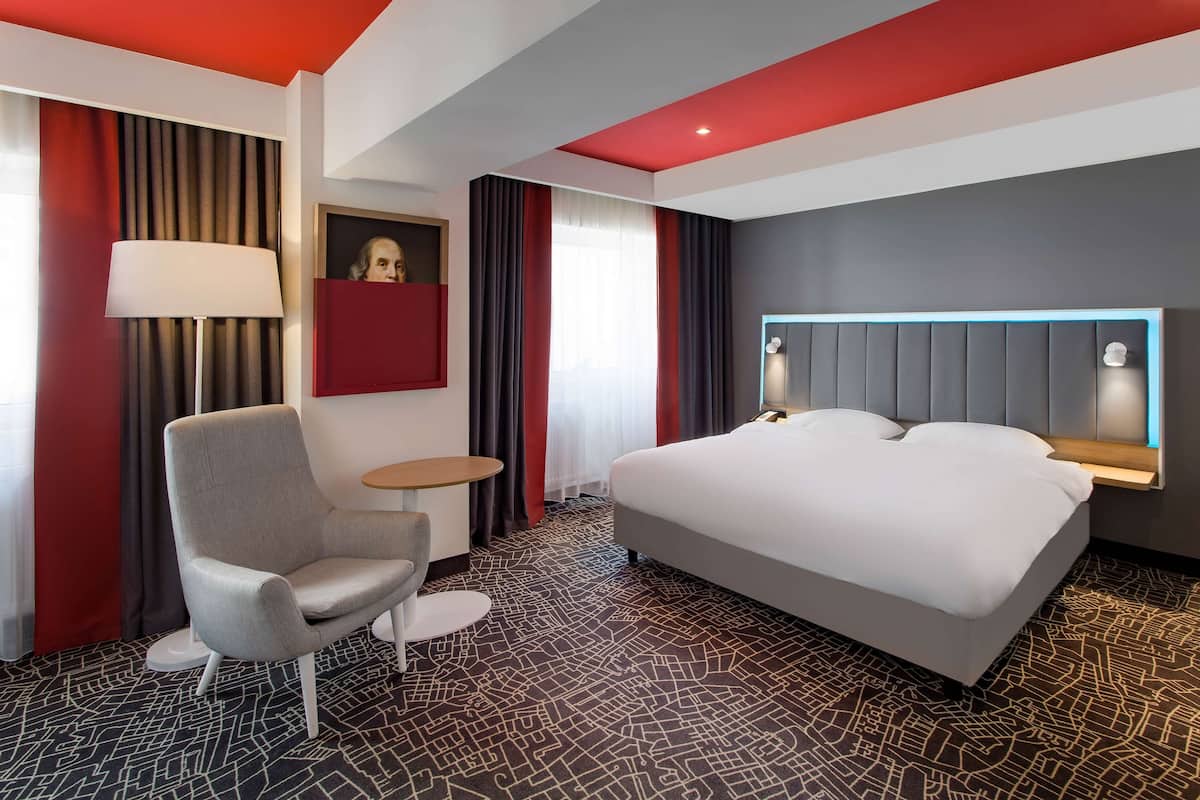 A hotel room with a large bed, a desk, a chair, a lamp, and a painting on the wall.