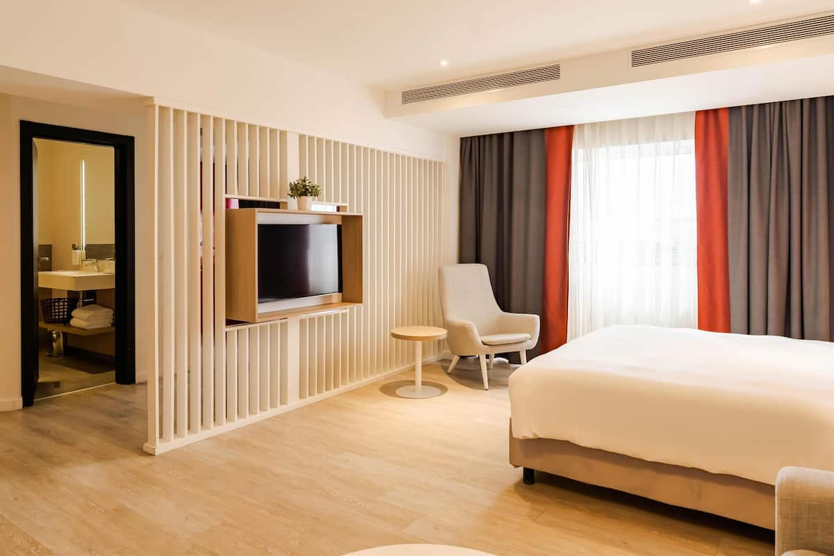 A hotel room with a large bed, a TV mounted on the wall, a small round table, a chair, and a wardrobe.