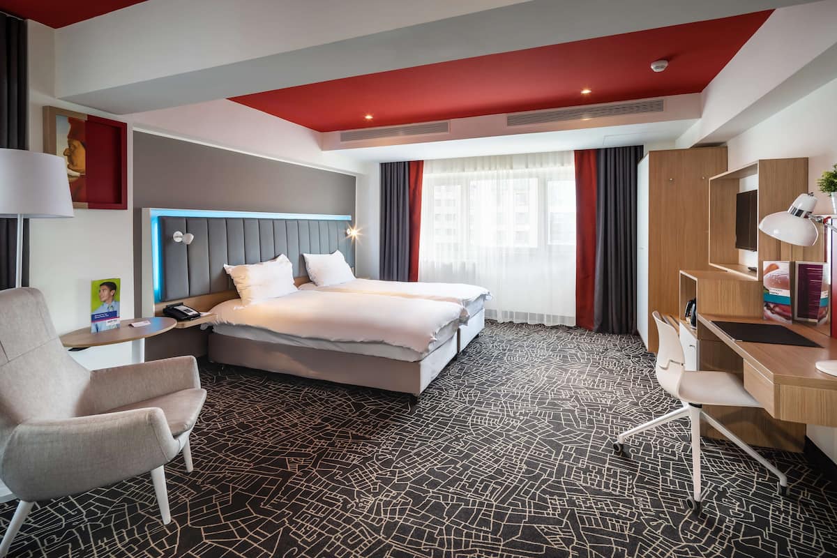 A hotel room with a bed, a desk, a chair, a lamp, and a window with curtains.