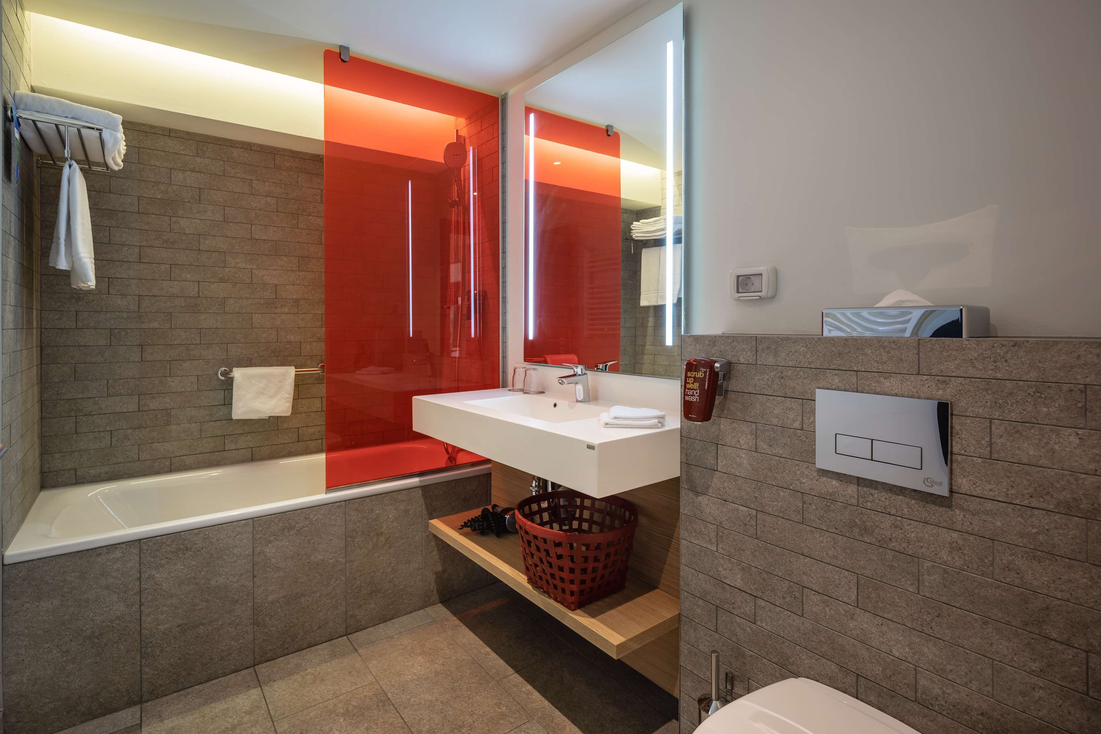 A modern bathroom with a white sink, a glass shower enclosure, and a red accent wall.
