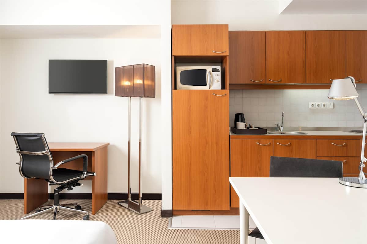 A modern hotel room with a wooden desk, a black office chair, a standing lamp, a microwave in a wooden cabinet, and a white table.