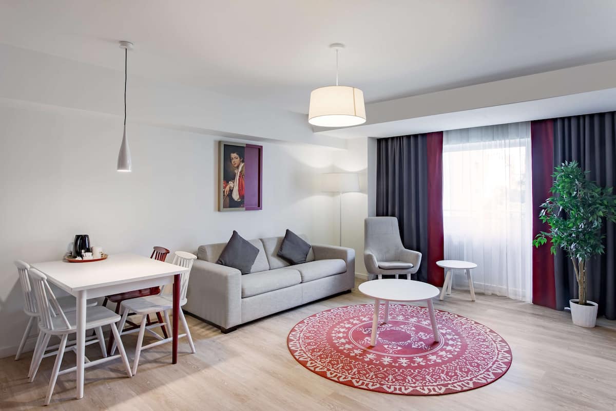 A modern living room with a white sofa, grey armchairs, a dining table with chairs, a red patterned rug, and a potted plant.