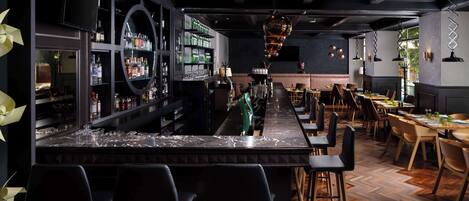 Zona de bar con encimera de mármol, taburetes negros y una barra larga. Se observan estantes con botellas y un televisor montado en la pared.