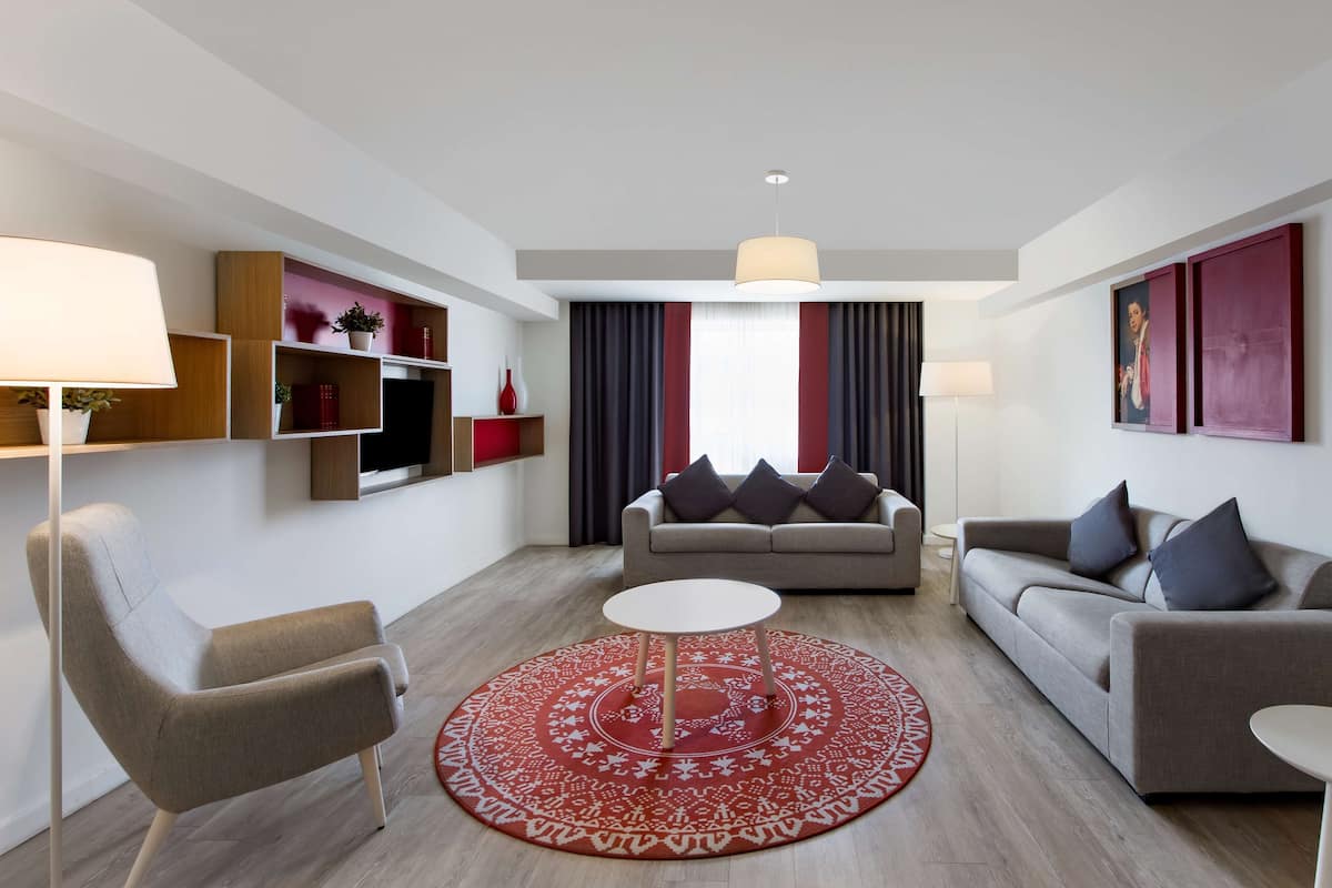A modern living room with a grey sofa, a round coffee table, and a red patterned rug. There are shelves with decorative items and a large window with curtains.