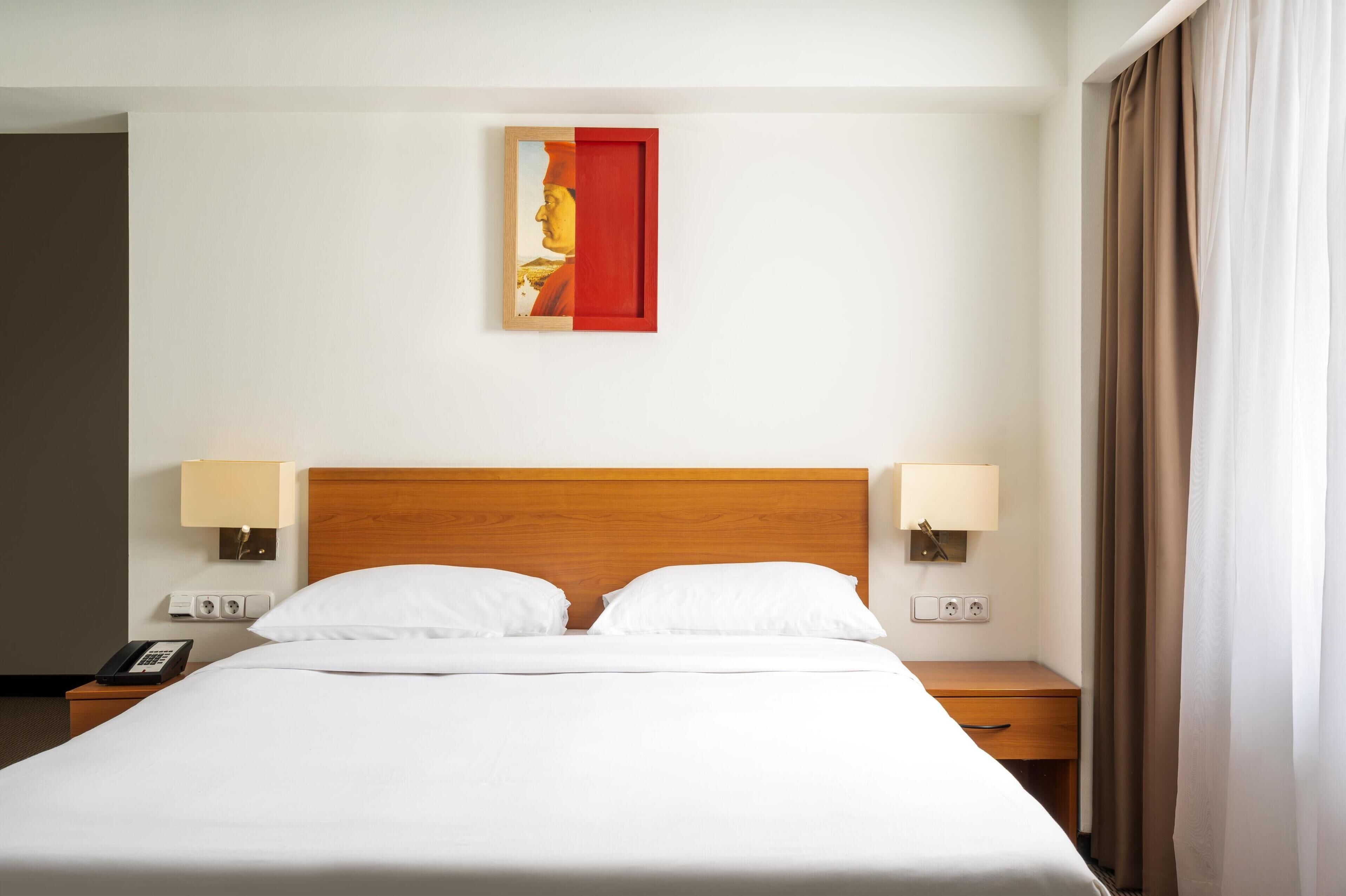 A hotel room with a wooden headboard, two bedside lamps, a telephone, and a painting on the wall.